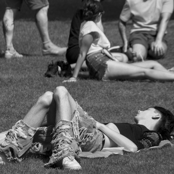 Black-and-white photograph of a person resting on grass in fringed boots and sunglasses, with other seated people behind them.