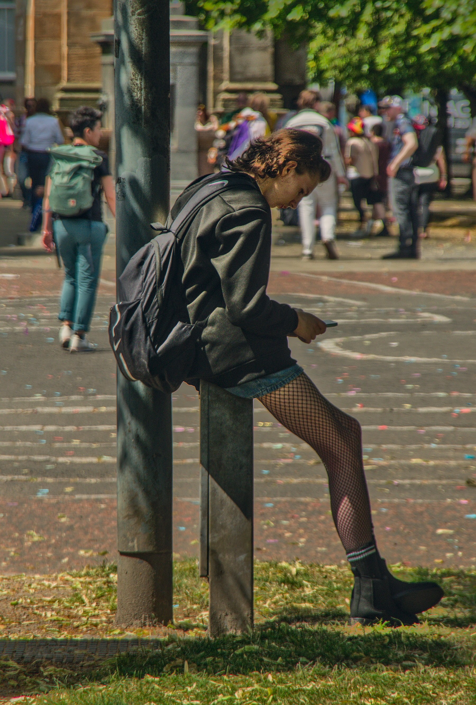 Person in a black jacket and fishnet tights sits on a metal bollard, looking at a phone, while Pride crowds gather behind them on Glasgow Green.