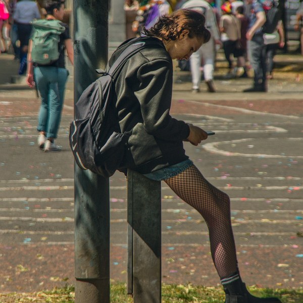 Person in a black jacket and fishnet tights sits on a metal bollard, looking at a phone, while Pride crowds gather behind them on Glasgow Green.