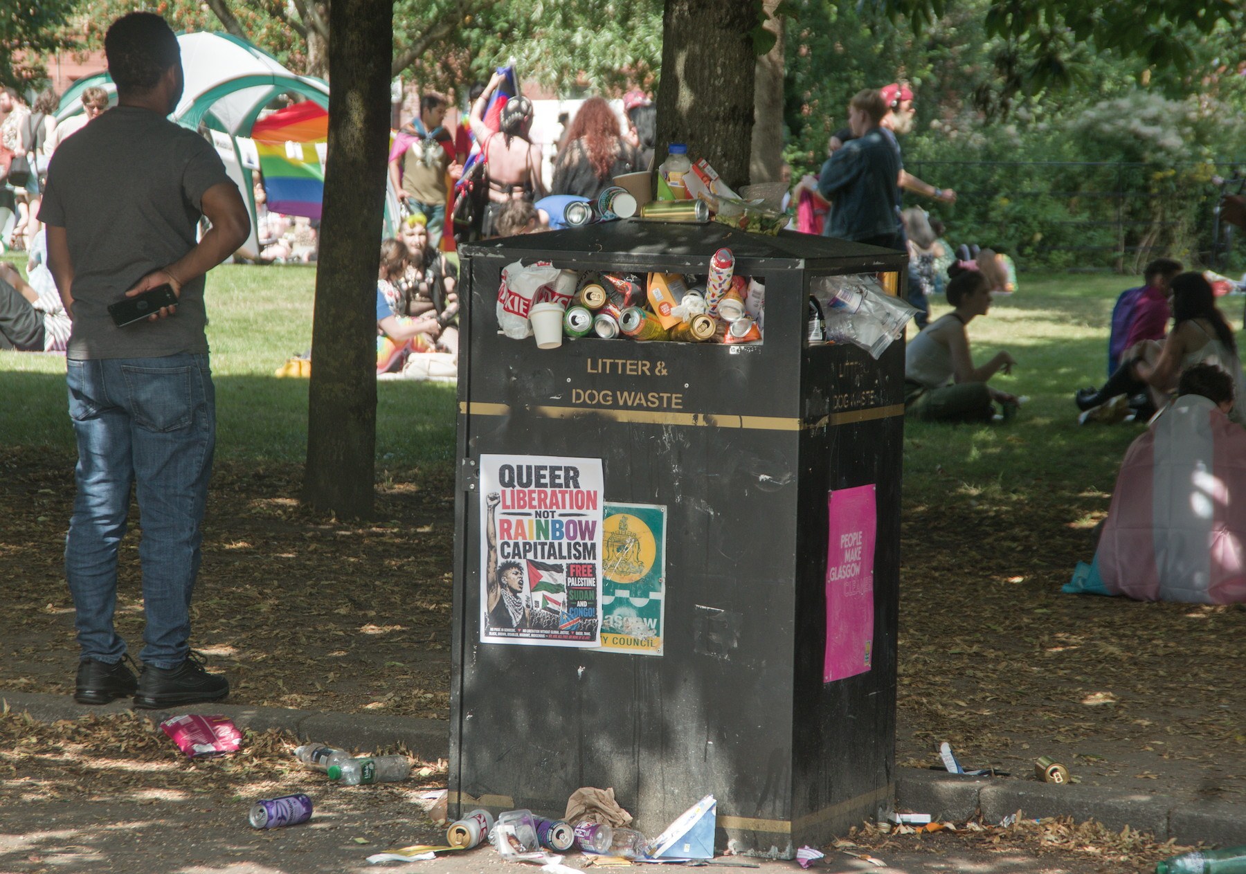 Overflowing litter and dog-waste bin beneath trees at Glasgow Green during Pride 2026, with people resting on the grass behind it.