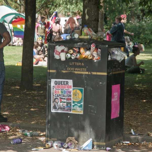 Overflowing litter and dog-waste bin beneath trees at Glasgow Green during Pride 2026, with people resting on the grass behind it.