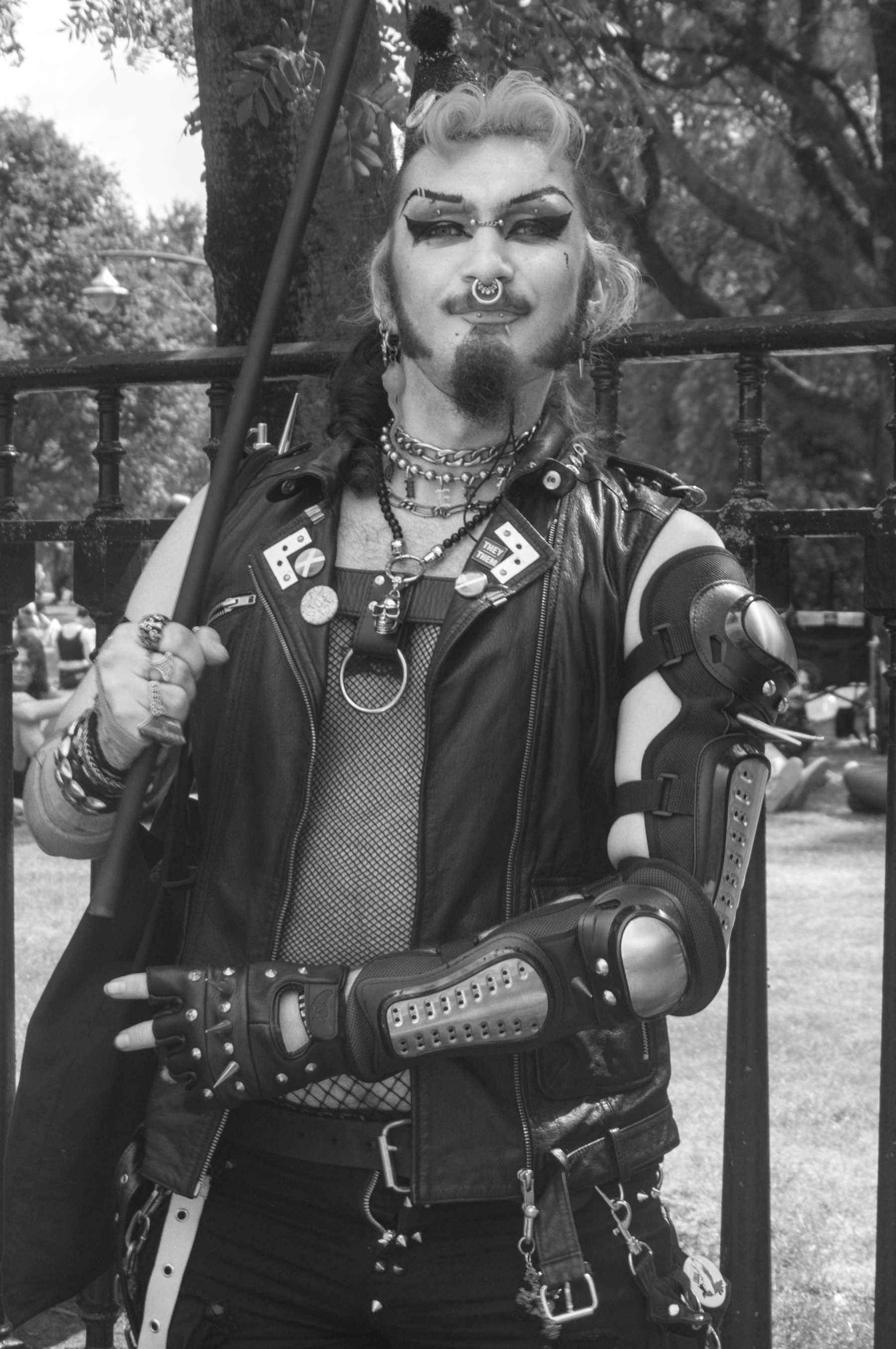 Black-and-white portrait of a person with dramatic eye makeup, a septum ring and facial hair, wearing a leather vest, layered chains, fishnet top and protective arm guards while holding a pole beside an iron fence.