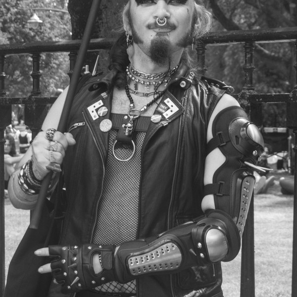 Black-and-white portrait of a person with dramatic eye makeup, a septum ring and facial hair, wearing a leather vest, layered chains, fishnet top and protective arm guards while holding a pole beside an iron fence.