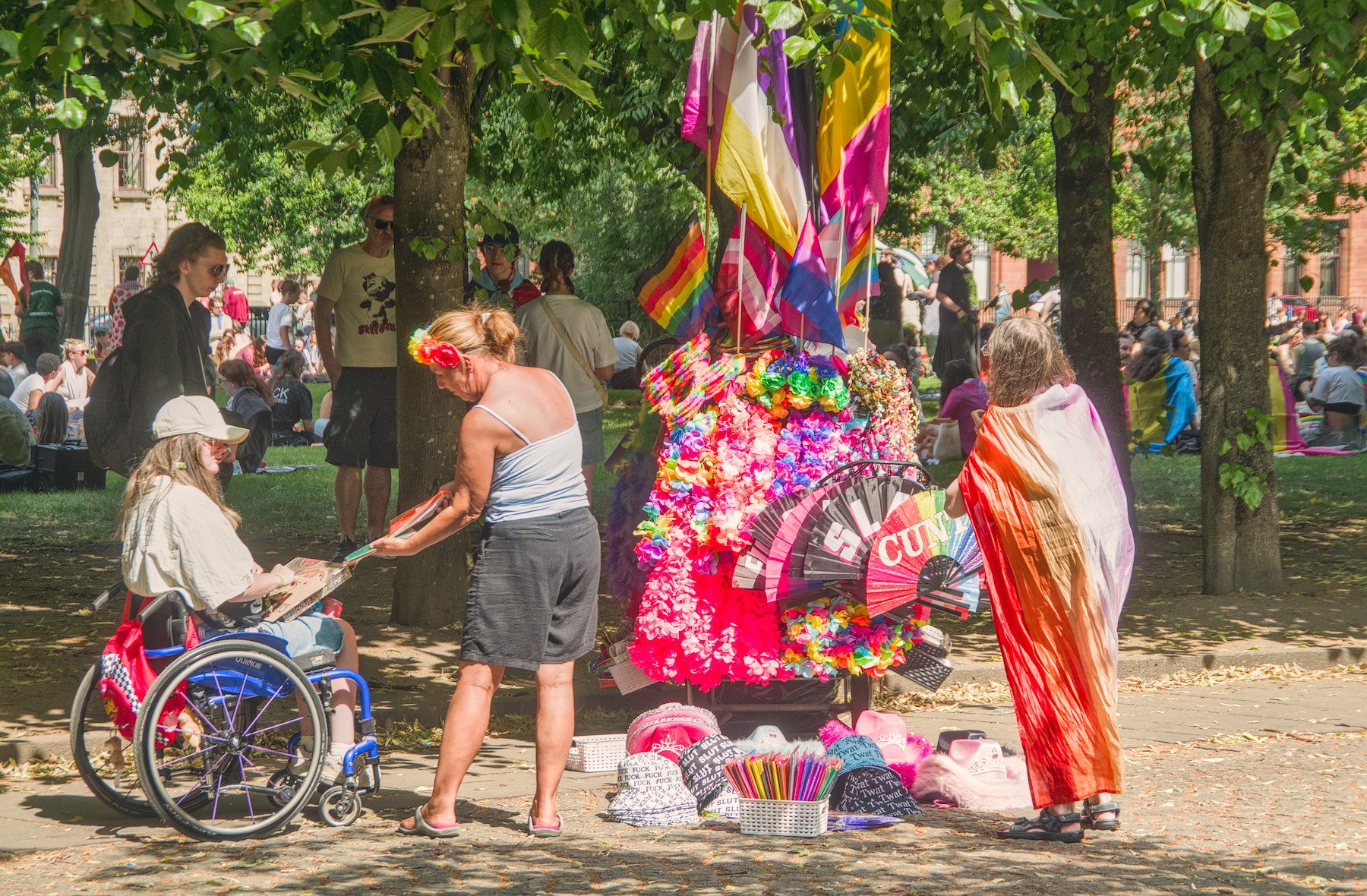 A wheelchair user and two standing people beside a brightly decorated Pride display covered in rainbow colours and flags beneath trees at Glasgow Green.