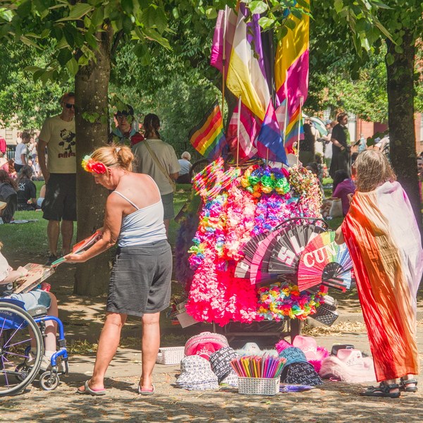 A wheelchair user and two standing people beside a brightly decorated Pride display covered in rainbow colours and flags beneath trees at Glasgow Green.