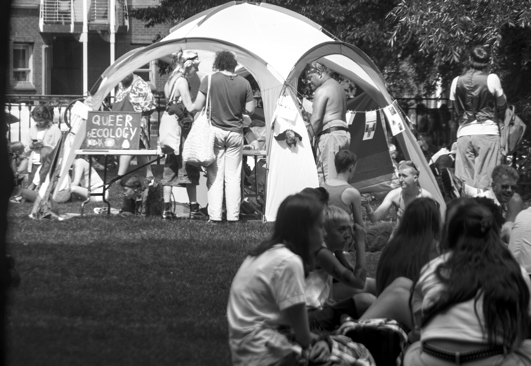 People gather beside a canopy tent with a sign reading “Queer Ecology” at Glasgow Green; seated attendees fill the foreground.