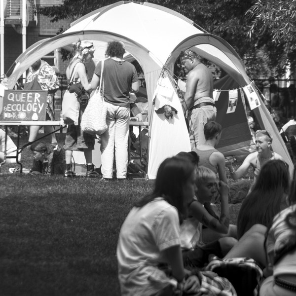 People gather beside a canopy tent with a sign reading “Queer Ecology” at Glasgow Green; seated attendees fill the foreground.