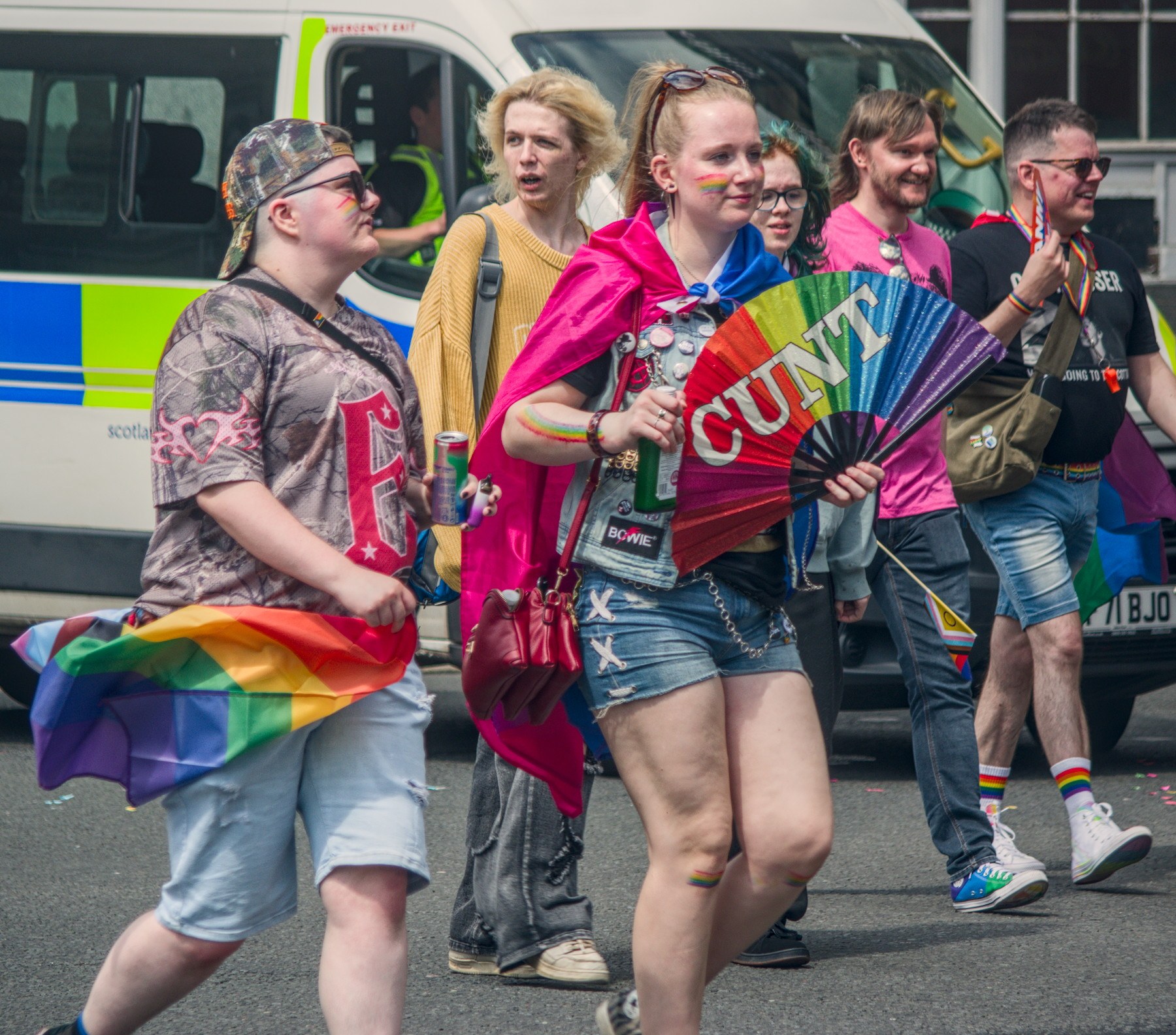 A group of Pride attendees walks along a street beside a police van. In front, a person wearing a bright pink cape carries a rainbow folding fan reading “CUNT”, while another person holds a rainbow flag.