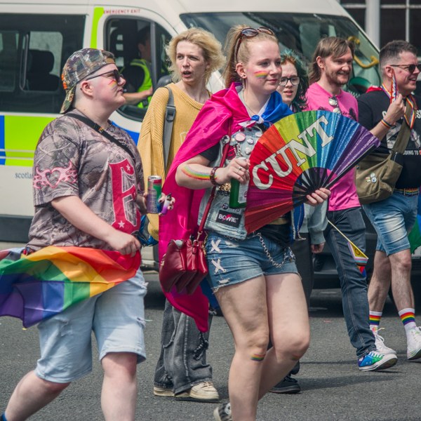 A group of Pride attendees walks along a street beside a police van. In front, a person wearing a bright pink cape carries a rainbow folding fan reading “CUNT”, while another person holds a rainbow flag.