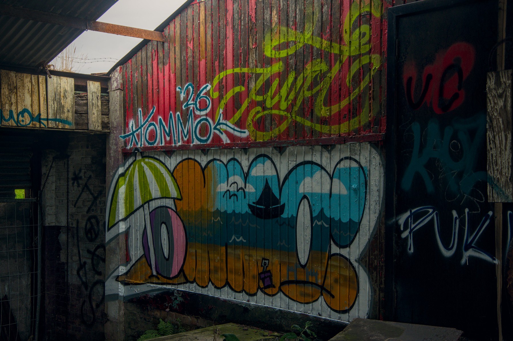 Colourful graffiti covers corrugated metal beneath red timber cladding inside a dilapidated roofed structure, beside a dark marked doorway.