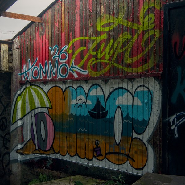Colourful graffiti covers corrugated metal beneath red timber cladding inside a dilapidated roofed structure, beside a dark marked doorway.