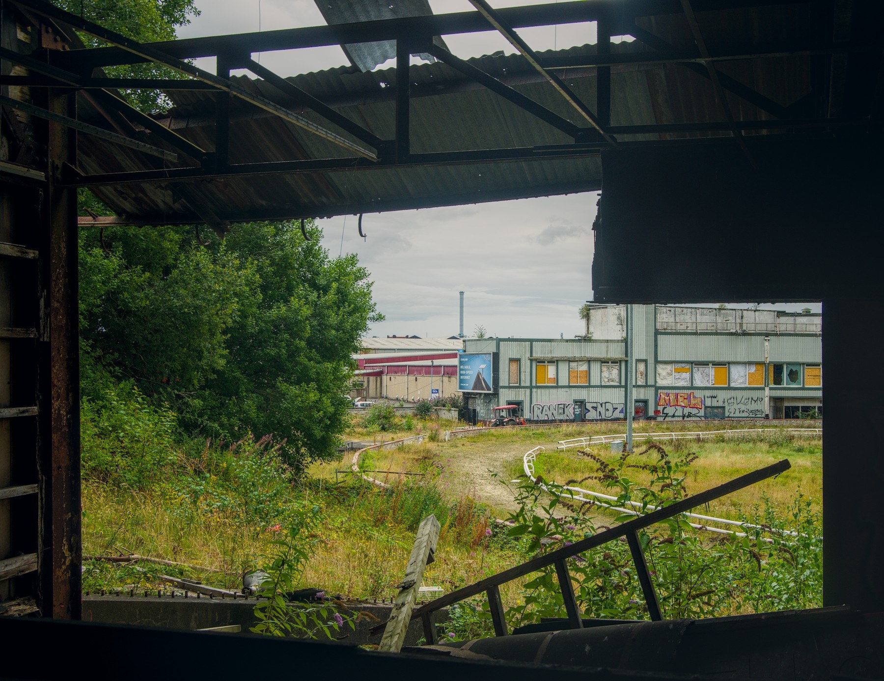 View through the dark steel frame of a roofless industrial structure towards overgrown ground and a low factory building with painted panels and wall markings.