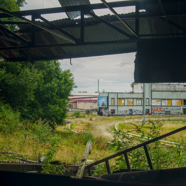 View through the dark steel frame of a roofless industrial structure towards overgrown ground and a low factory building with painted panels and wall markings.