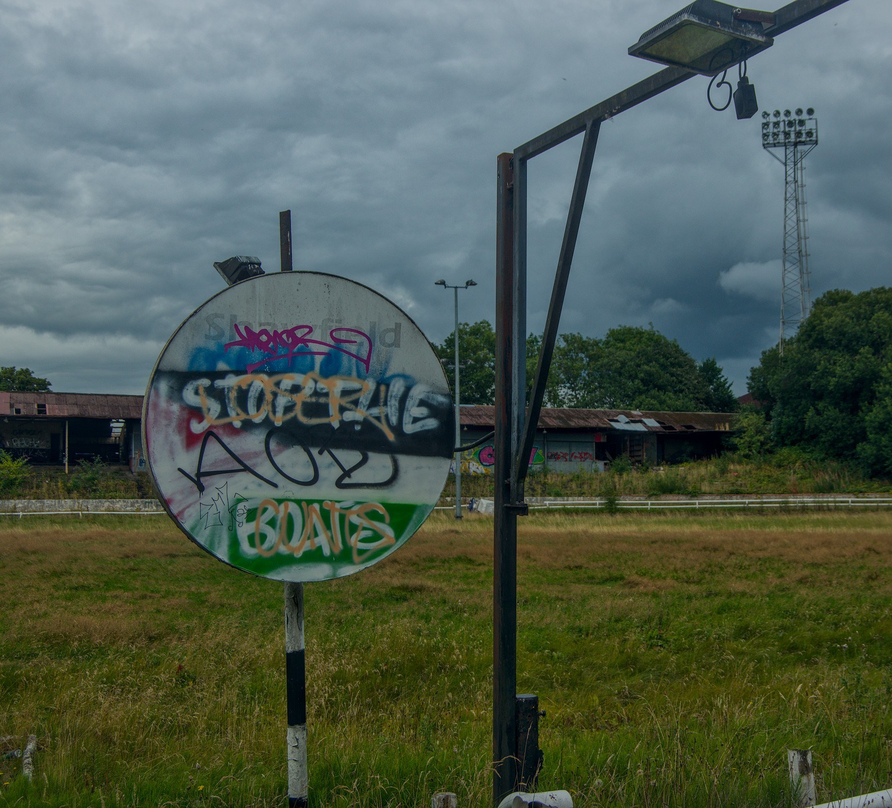 Spray-painted circular sign beside a metal support and floodlight tower in overgrown grounds at Shawfield Track under heavy clouds.