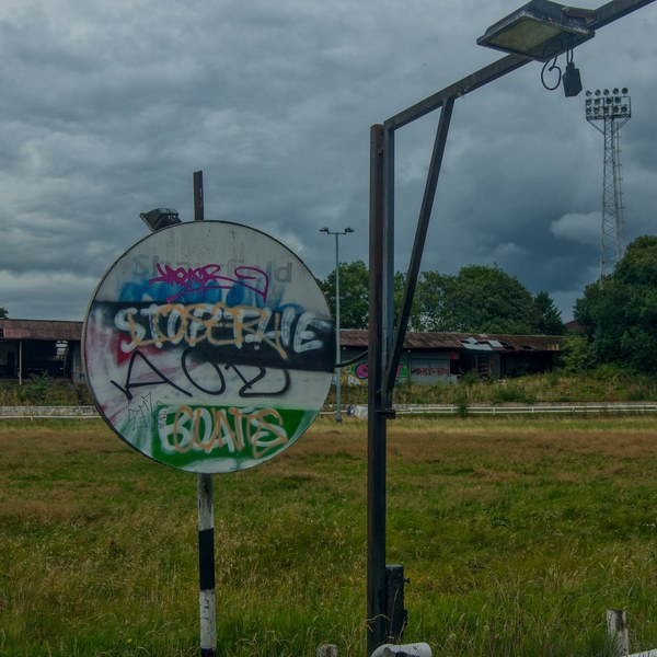 Spray-painted circular sign beside a metal support and floodlight tower in overgrown grounds at Shawfield Track under heavy clouds.