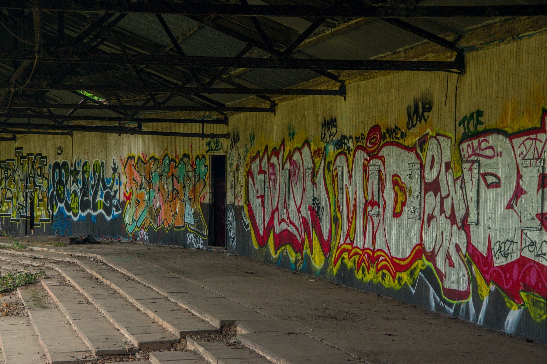 Interior of a disused corrugated-metal shelter, with a long wall covered in multicoloured graffiti, exposed roof trusses, cracked concrete flooring and stepped seating at left.