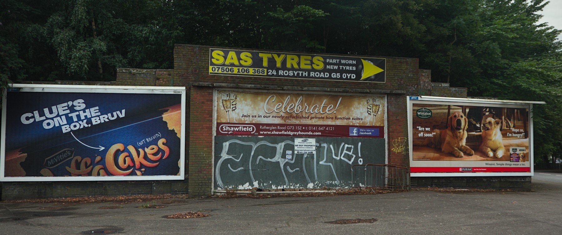 Brick roadside frontage in Shawfield with SAS Tyres, Cakes and dog-food advertisements above a shutter covered in white lettering.