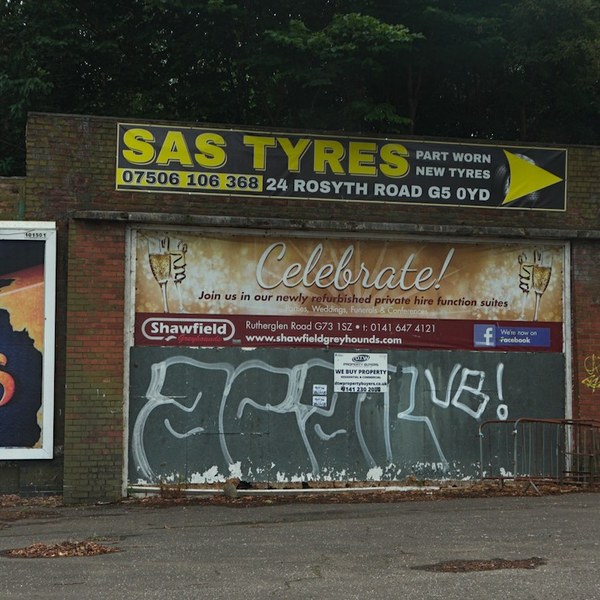 Brick roadside frontage in Shawfield with SAS Tyres, Cakes and dog-food advertisements above a shutter covered in white lettering.