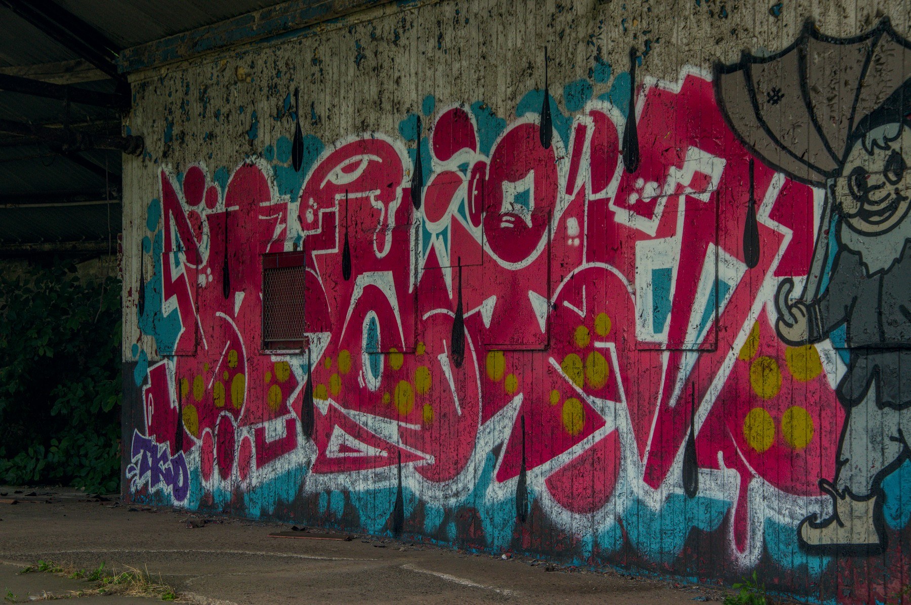 Large red, white and turquoise graffiti lettering with lime-green dots covers a weathered concrete wall beside a small brown vent, under a dark roof overhang.