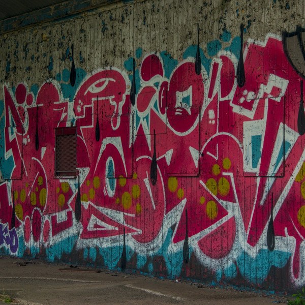 Large red, white and turquoise graffiti lettering with lime-green dots covers a weathered concrete wall beside a small brown vent, under a dark roof overhang.