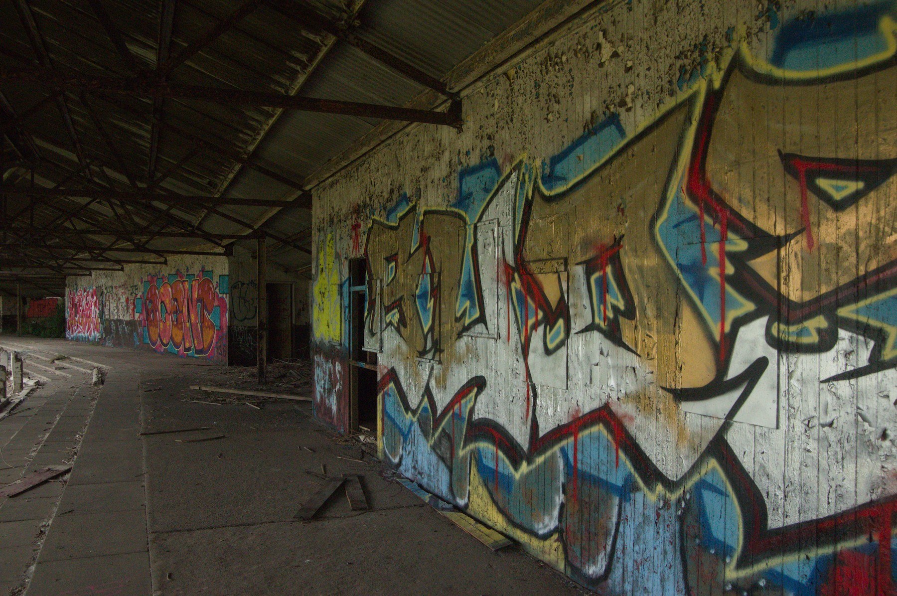 Large multicoloured graffiti pieces cover a weathered concrete wall beside a curving paved walkway under a corrugated canopy at Shawfield Track.
