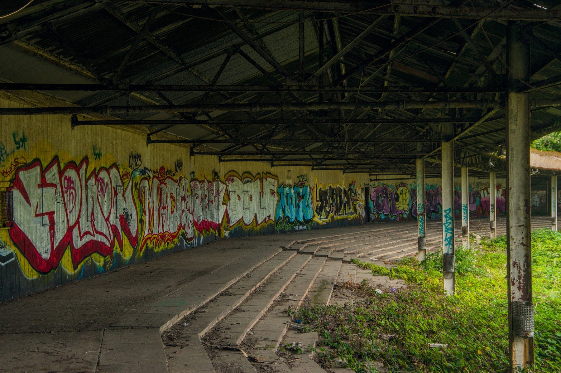 Graffiti-covered wall beneath a dark metal shelter, with curved concrete terrace steps and weeds growing beside support columns at Shawfield Track.