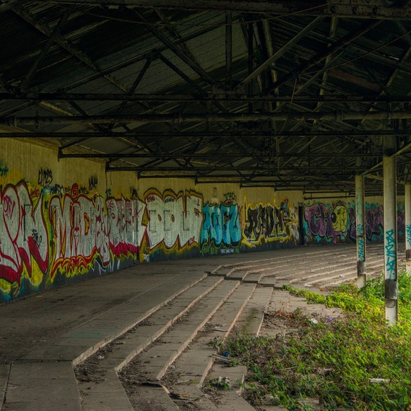 Graffiti-covered wall beneath a dark metal shelter, with curved concrete terrace steps and weeds growing beside support columns at Shawfield Track.