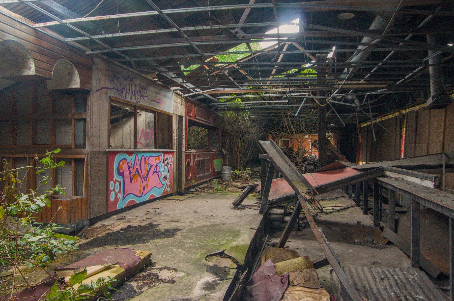 Graffiti-covered wooden service stalls and broken counters inside a derelict structure with a damaged corrugated roof.