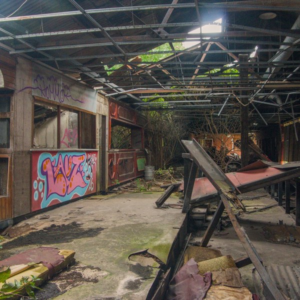 Graffiti-covered wooden service stalls and broken counters inside a derelict structure with a damaged corrugated roof.