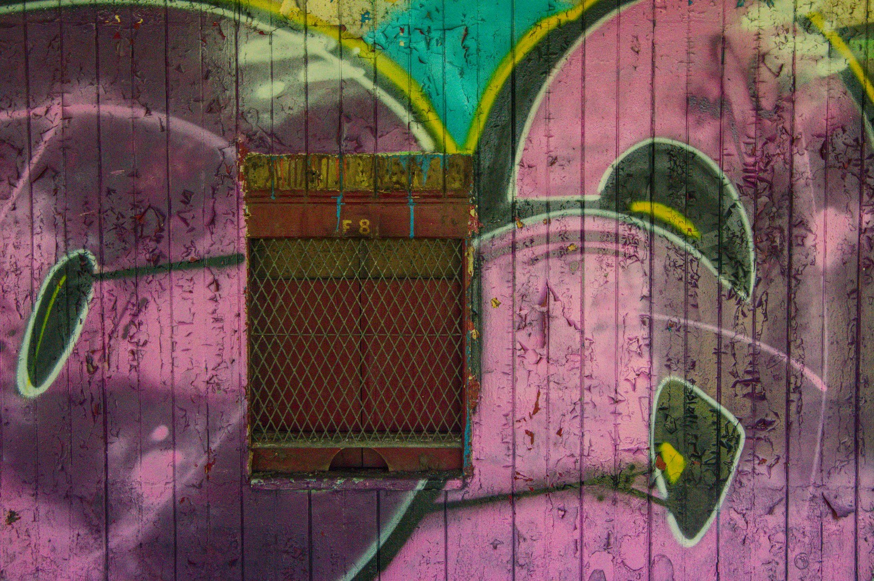 Rusty metal mesh vent marked F8 set into a weathered pink, turquoise and black graffiti mural on a wooden wall.