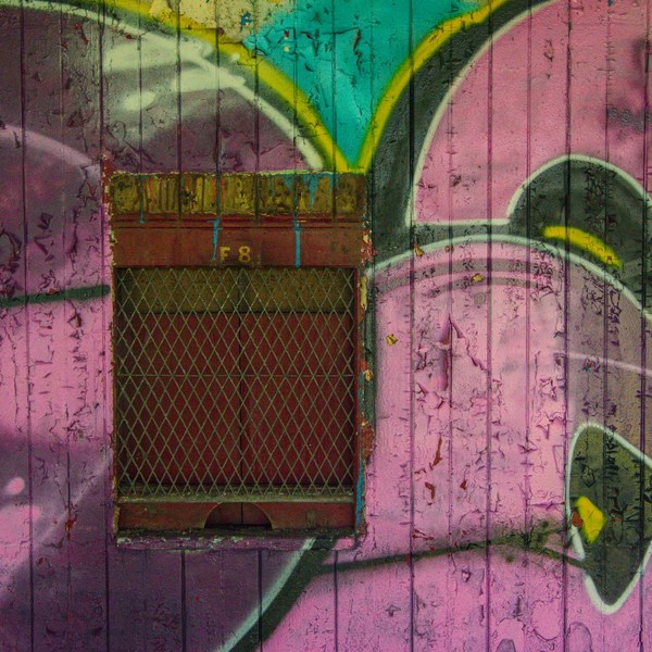 Rusty metal mesh vent marked F8 set into a weathered pink, turquoise and black graffiti mural on a wooden wall.