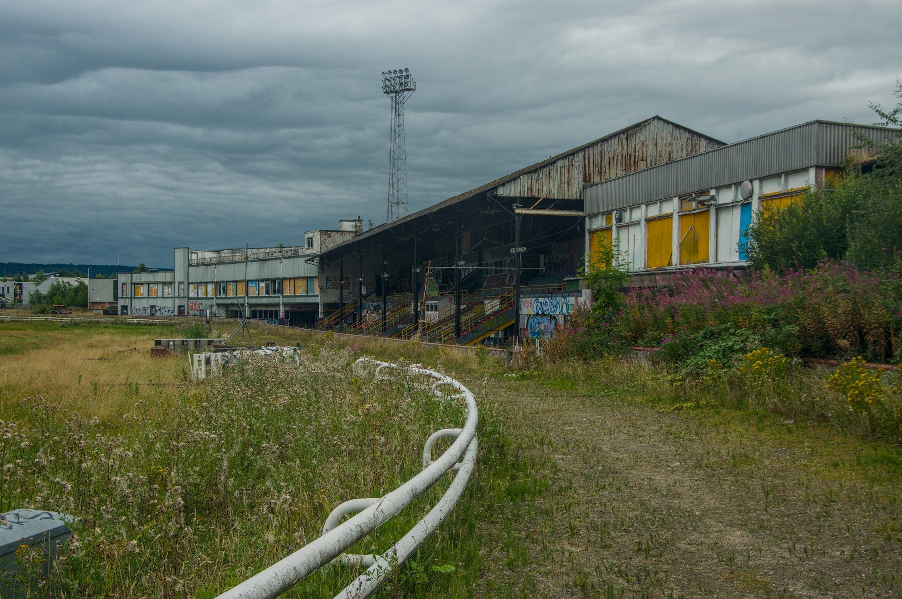 Curving white track rail beside overgrown grass, leading towards a weathered grandstand with yellow-covered windows under heavy clouds.