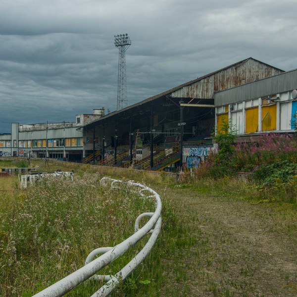 Curving white track rail beside overgrown grass, leading towards a weathered grandstand with yellow-covered windows under heavy clouds.