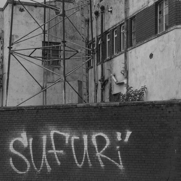 Black-and-white view of metal scaffolding against a weathered industrial building, above a dark brick boundary marked with large graffiti lettering.