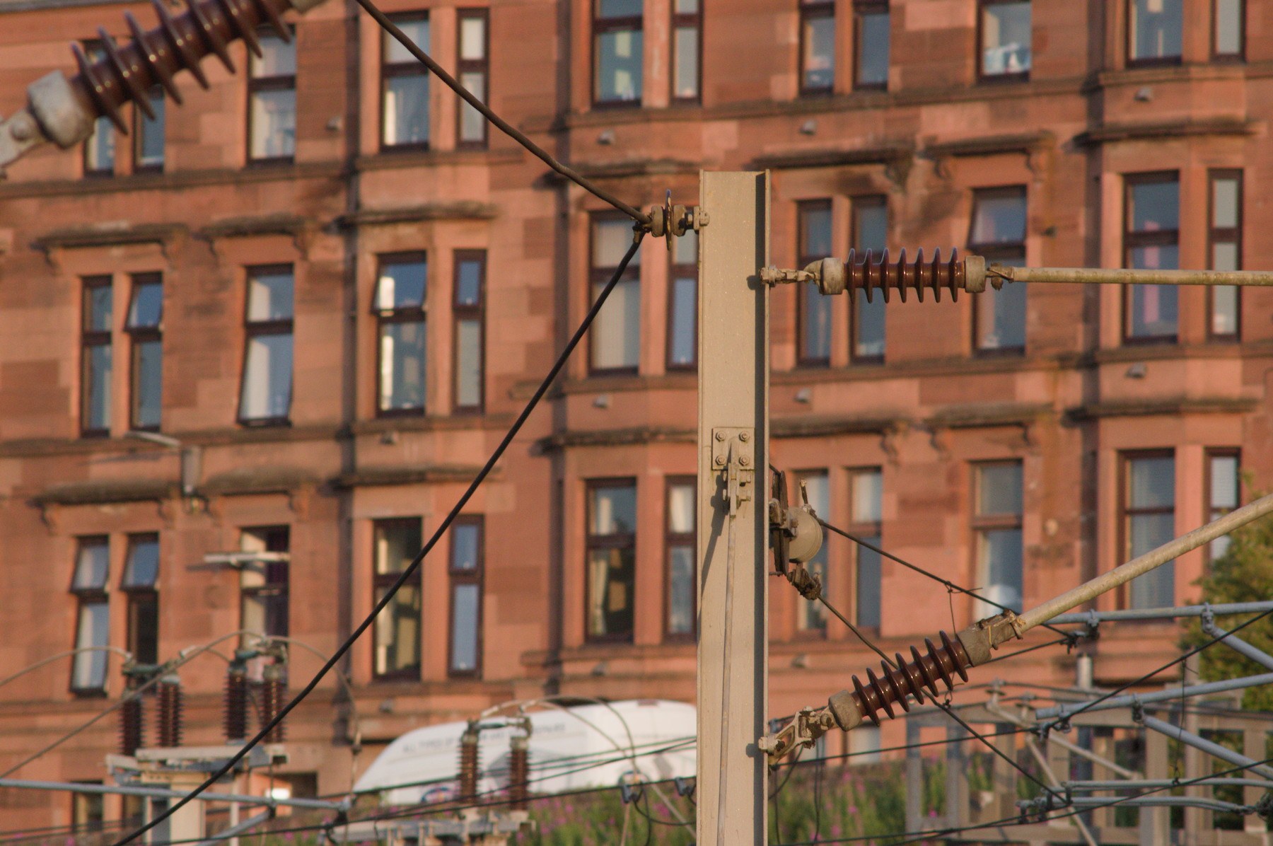 Railway overhead-line insulators and cables in front of a sunlit red sandstone building with rows of tall windows in Rutherglen.