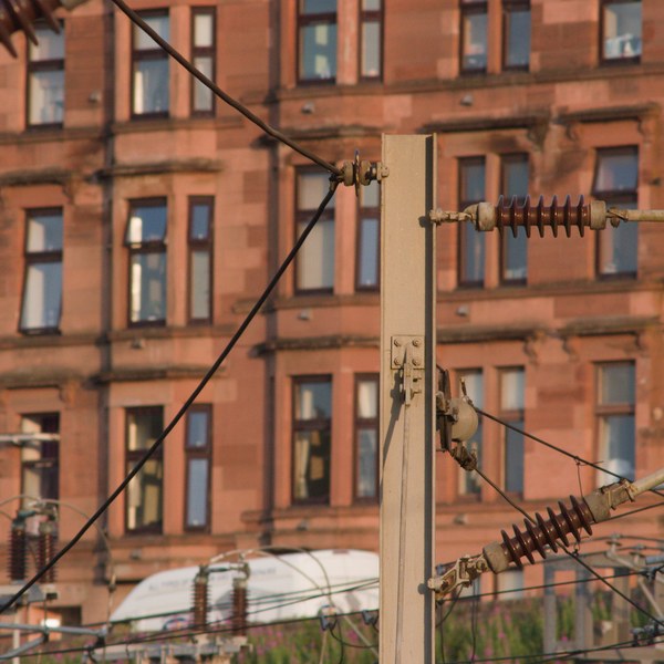 Railway overhead-line insulators and cables in front of a sunlit red sandstone building with rows of tall windows in Rutherglen.