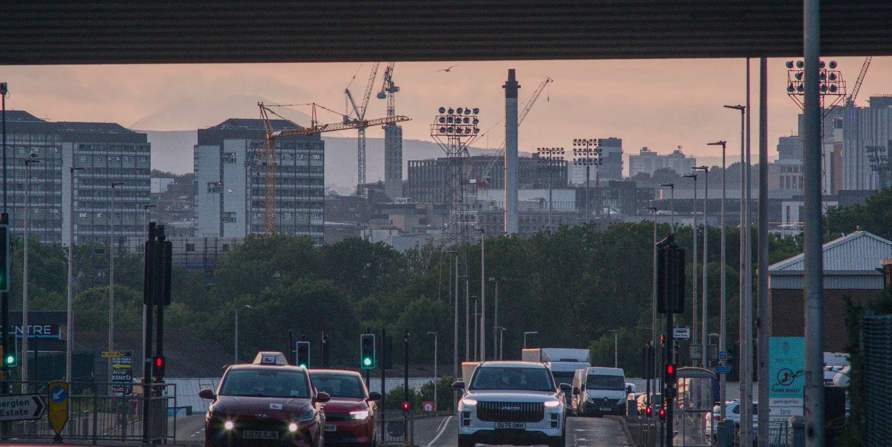 Evening traffic on a wet road in Rutherglen, viewed beneath an overpass toward distant cranes, floodlights and city buildings.