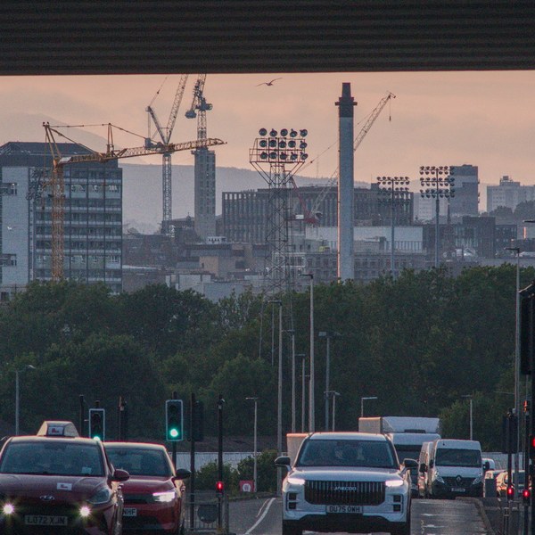 Evening traffic on a wet road in Rutherglen, viewed beneath an overpass toward distant cranes, floodlights and city buildings.