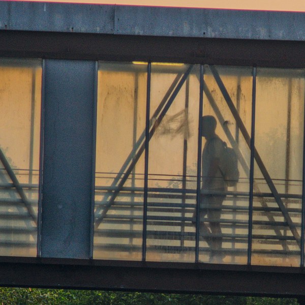 Silhouetted person walking through a glass-sided pedestrian bridge, framed by dark steel supports and warm evening light.