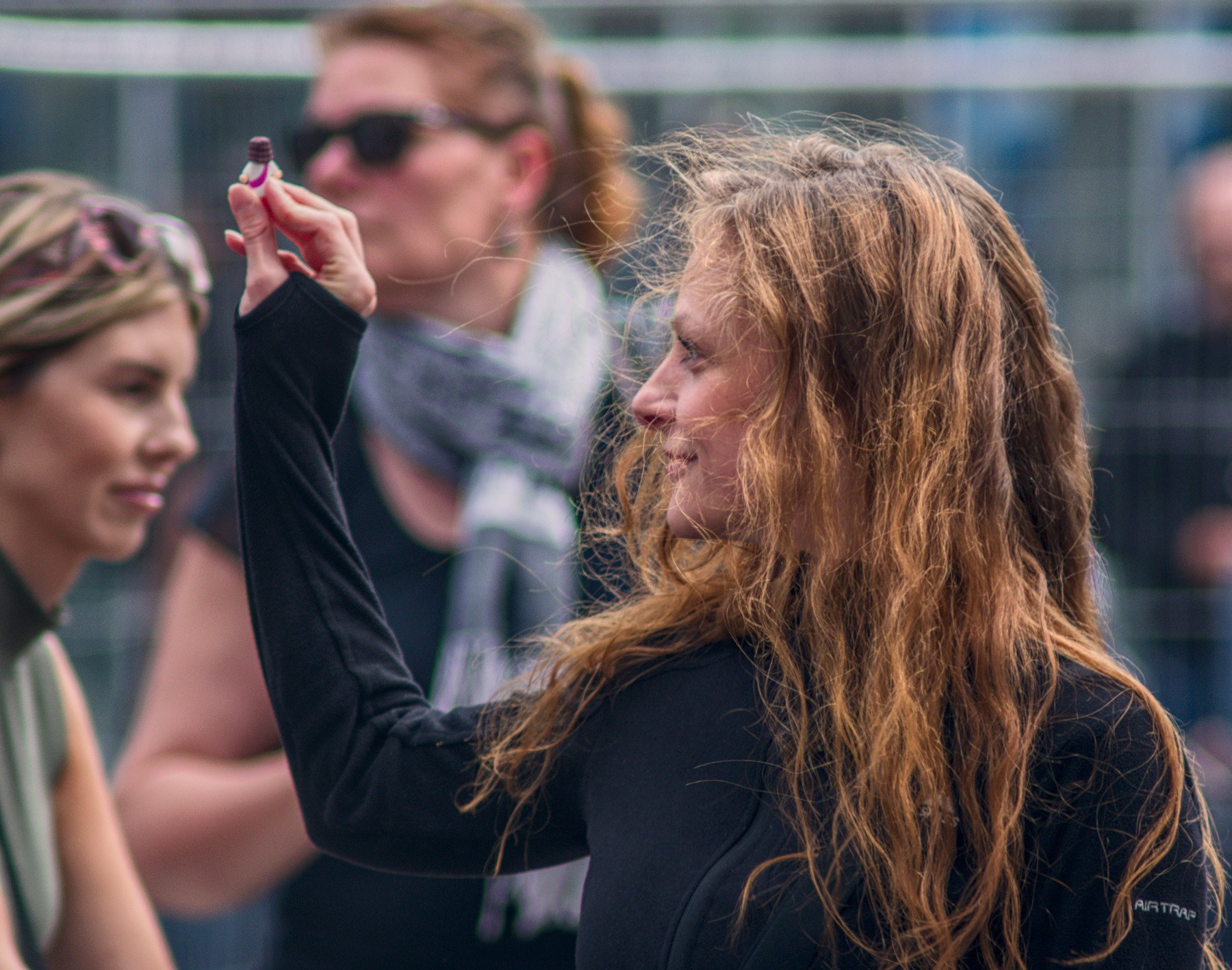 Side-profile close-up of a long-haired woman in black raising a small object between her fingers, with two women softly blurred behind her.