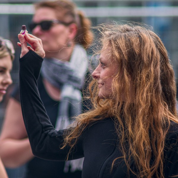 Side-profile close-up of a long-haired woman in black raising a small object between her fingers, with two women softly blurred behind her.