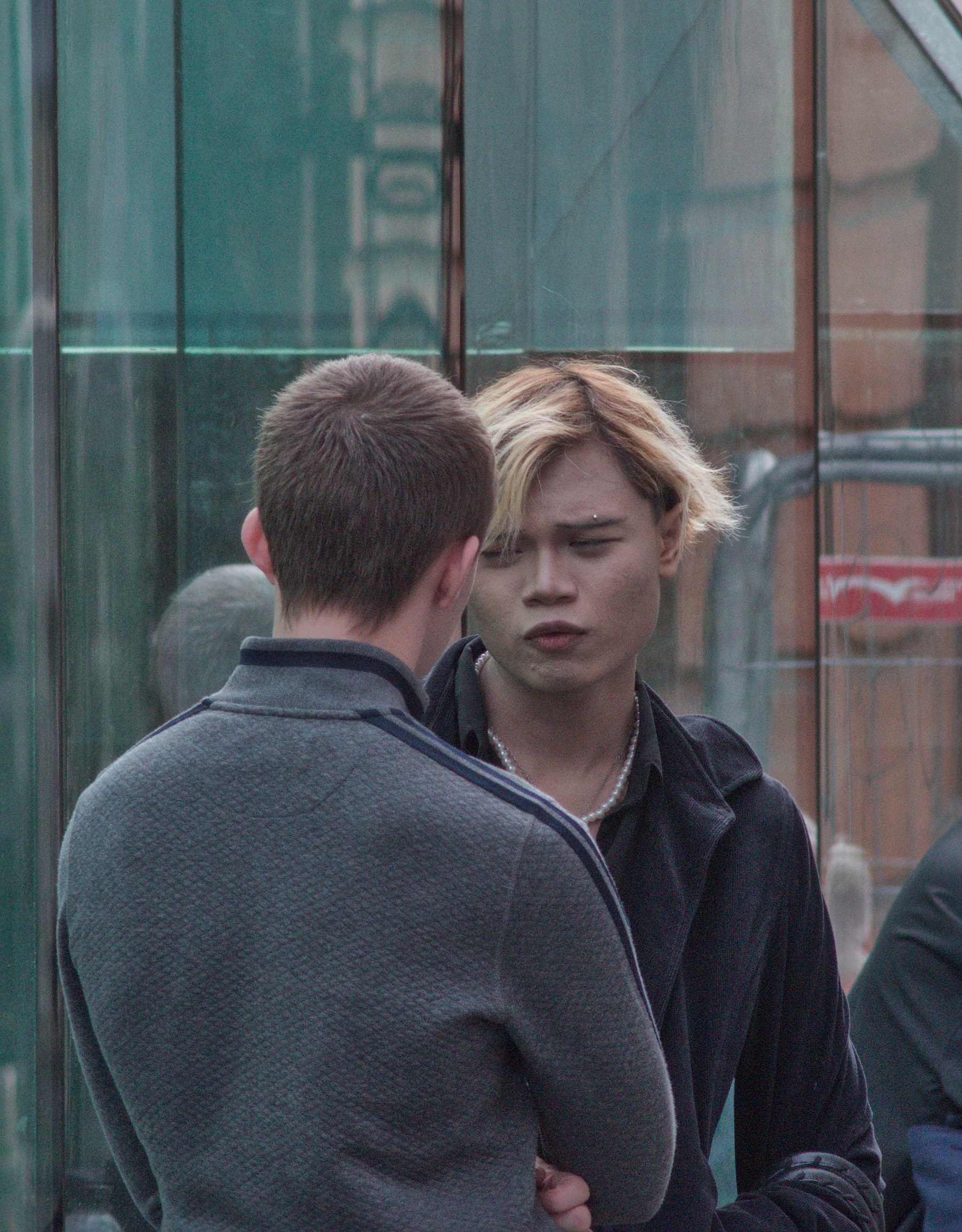 Two people stand close together in front of a reflective glass wall; one is seen from behind in a grey jacket while the other, with light-streaked hair and a dark coat, faces the camera.