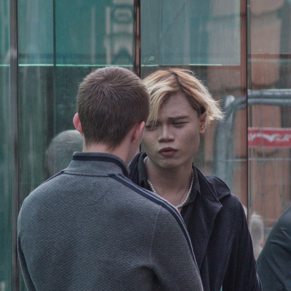 Two people stand close together in front of a reflective glass wall; one is seen from behind in a grey jacket while the other, with light-streaked hair and a dark coat, faces the camera.