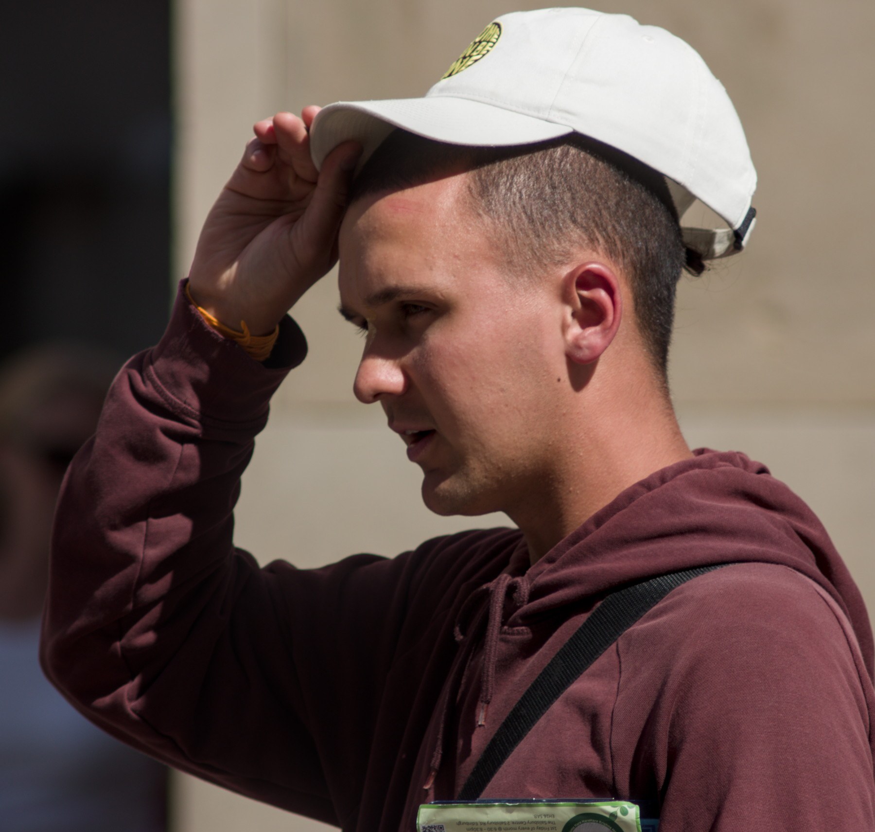 Side-profile portrait of a man in a burgundy hoodie adjusting a white baseball cap in bright sunlight.