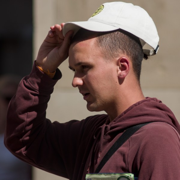 Side-profile portrait of a man in a burgundy hoodie adjusting a white baseball cap in bright sunlight.