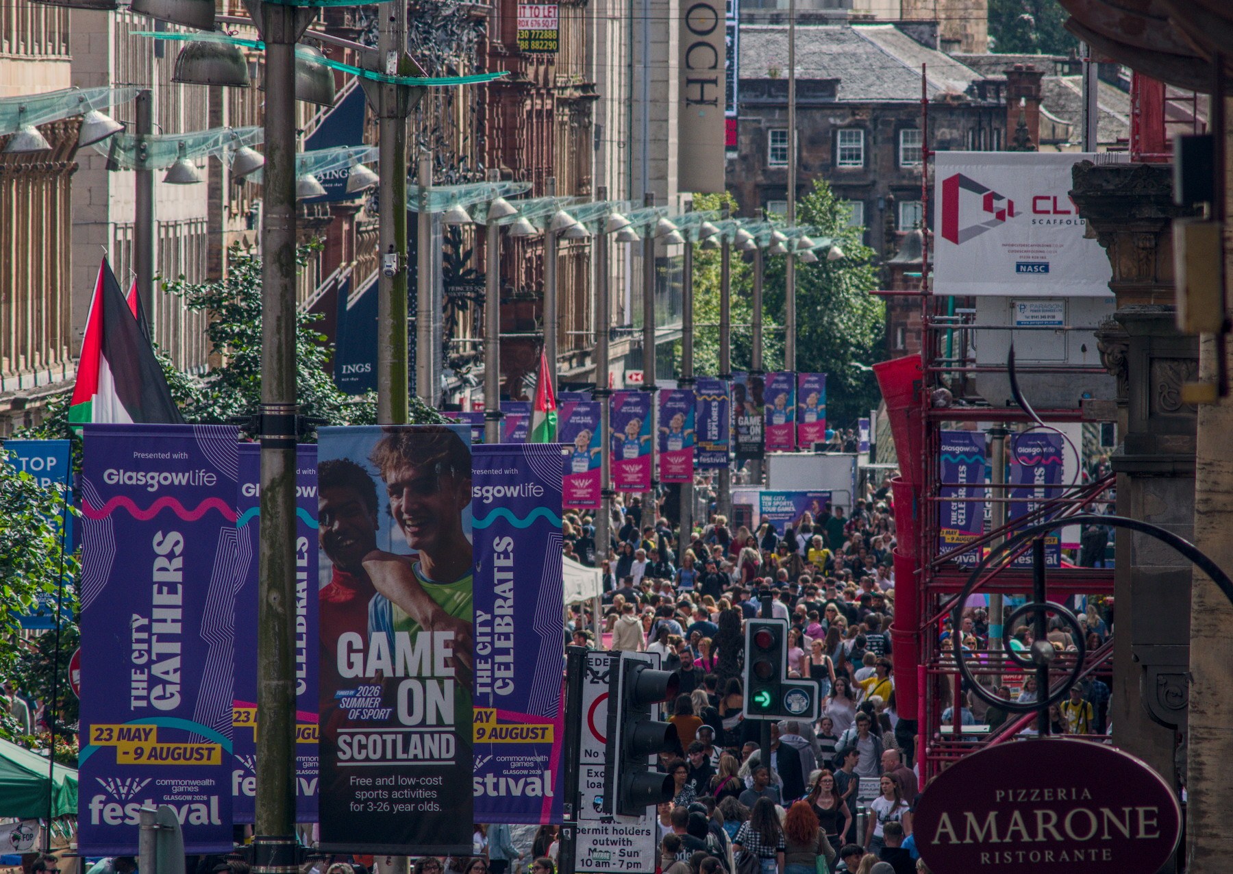 Telephoto view down a busy Glasgow city-centre street, with dense crowds beneath rows of hanging lights and colourful festival banners; traffic lights and shop signs frame the scene.