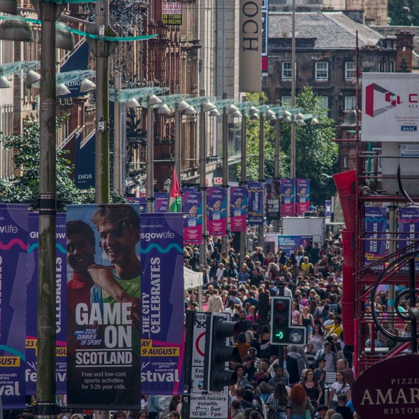 Telephoto view down a busy Glasgow city-centre street, with dense crowds beneath rows of hanging lights and colourful festival banners; traffic lights and shop signs frame the scene.