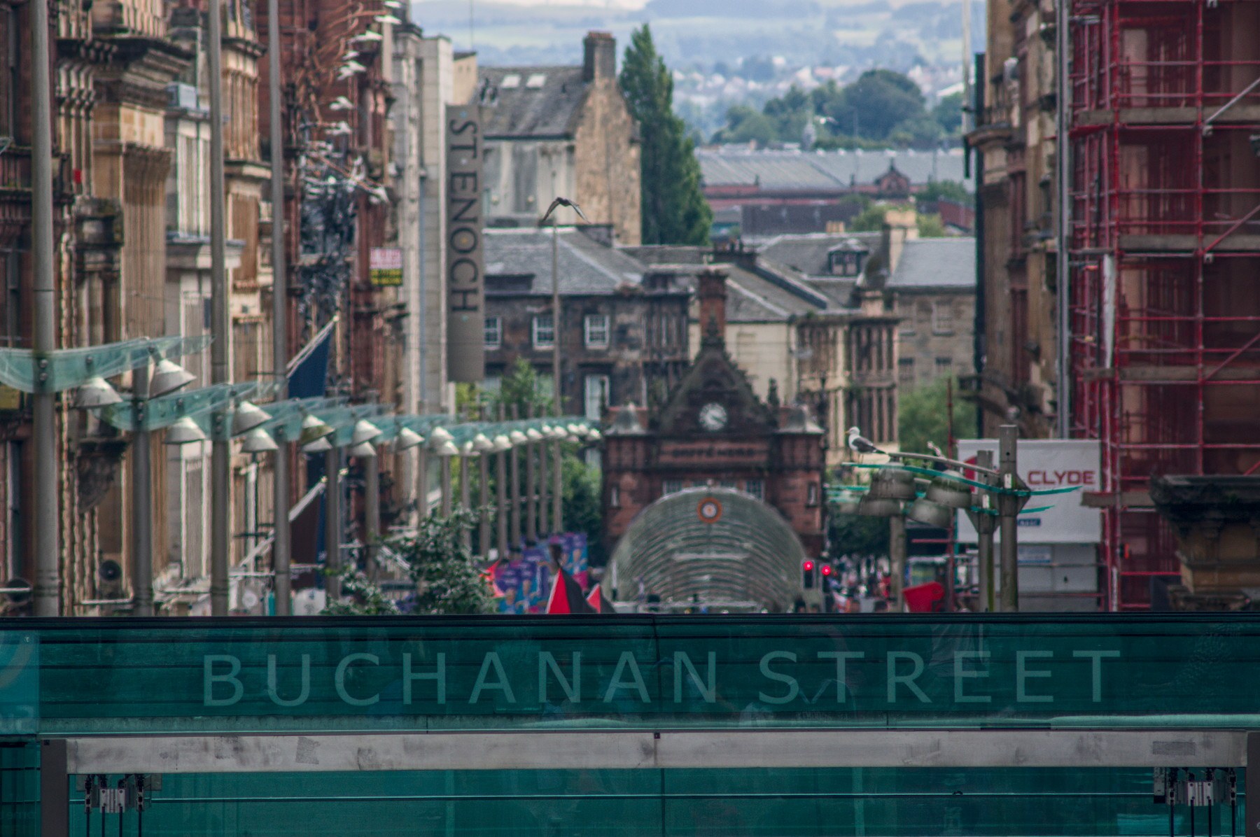 Telephoto view down Buchanan Street in Glasgow city centre, with historic stone buildings, shop signs and a green construction barrier in the foreground.