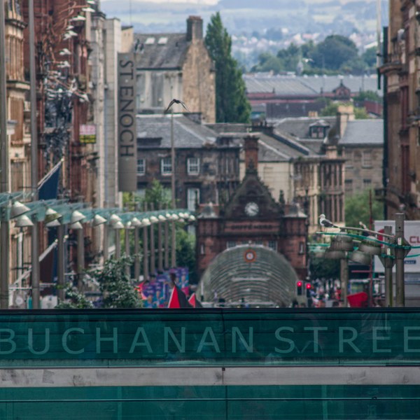 Telephoto view down Buchanan Street in Glasgow city centre, with historic stone buildings, shop signs and a green construction barrier in the foreground.