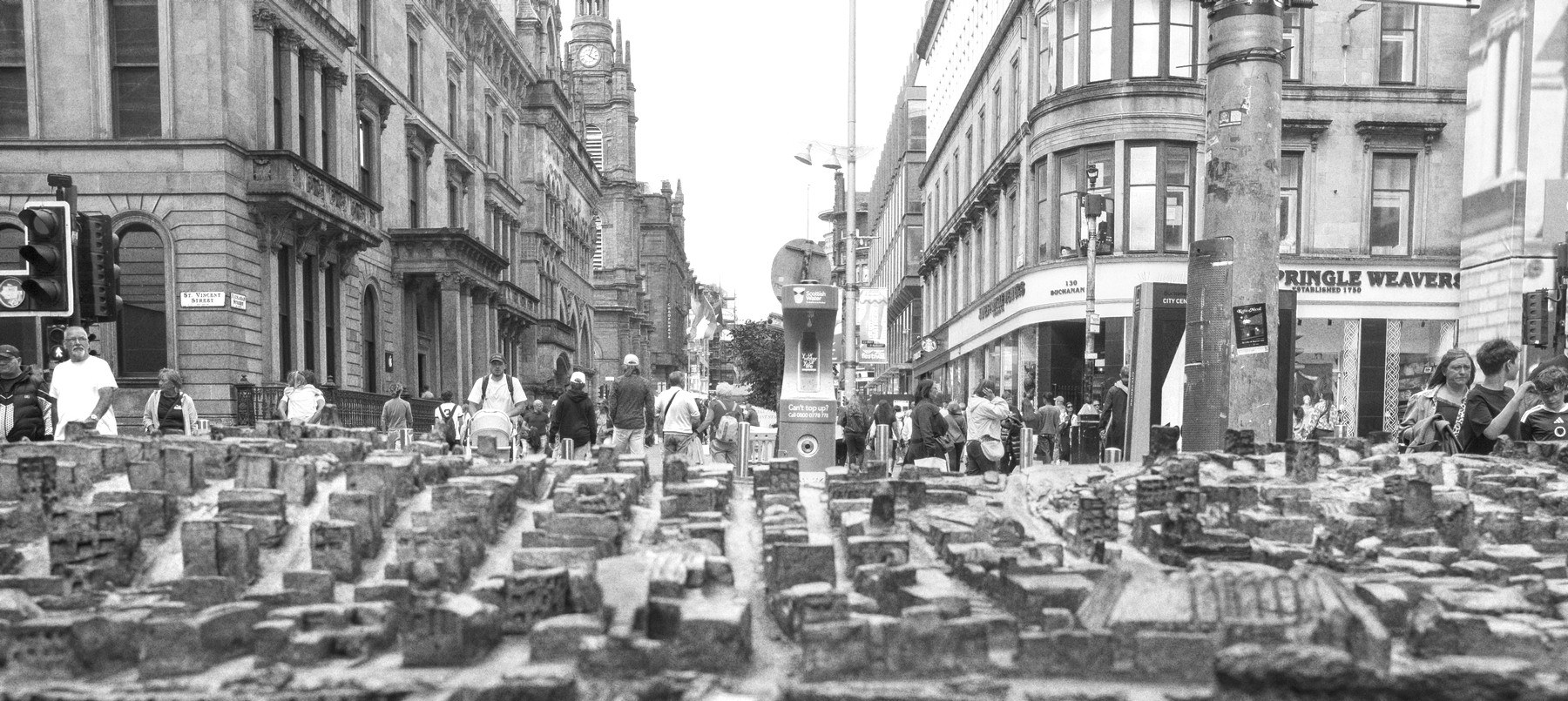 Black-and-white street scene in Glasgow city centre, with a table of densely arranged miniature building models in the foreground and pedestrians walking between tall historic commercial buildings behind.